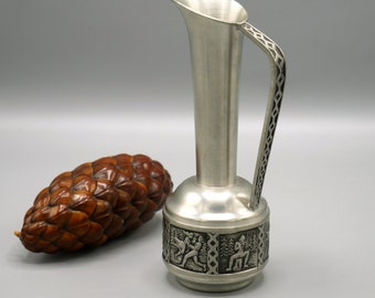 May include: A small pewter vase with a decorative handle and a design of figures and trees around the base.