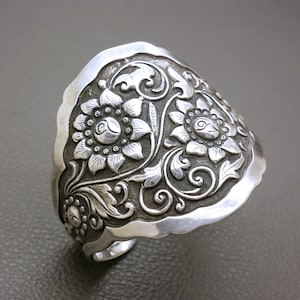 Op de afbeelding: Zilveren manchetarmband met een bloemenontwerp. De armband heeft gedetailleerde gravures van bloemen en bladeren. De randen hebben een geschulpte vormgeving. De manchet is gemaakt van glanzend, zilverkleurig metaal.
