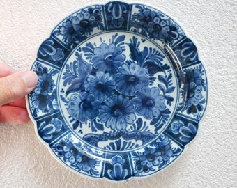 May include: A round ceramic plate with a scalloped edge, featuring a dark blue and white floral pattern. The center displays a cluster of stylized flowers, while the border is divided into segments containing additional floral motifs.
