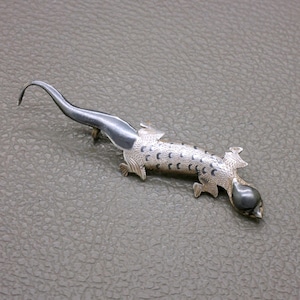 Op de afbeelding: Een zilverkleurige hagedisbroche met een gladde, donker metalen kop en staart. Het lichaam is getextureerd met een fijn kruisarceringpatroon en versierd met kleine, donkere, halvemaanvormige markeringen langs de rug en de zijkanten.