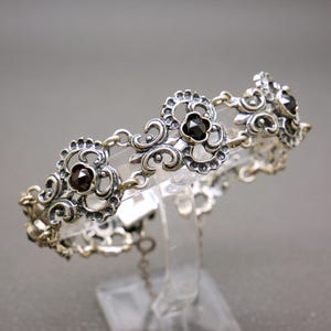 Könnte beinhalten: Silberfarbenes Armband mit kunstvollen floralen und Rankenmustern. Das Armband ist mit dunkelroten und schwarzen Edelsteinen verziert, die in die floralen Elemente eingesetzt sind. Die filigranen Details und Kontrastfarben erzeugen eine Vintage-Ästhetik.