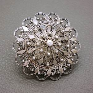 May include: A silver filigree brooch with a round design and a pearl in the centre.