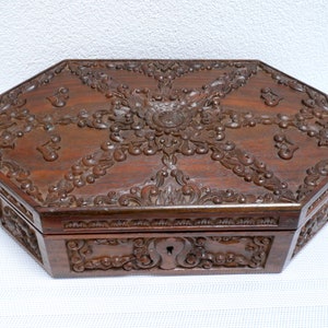 以下が含まれることがあります： 濃い茶色の八角形の木箱で、複雑な花の彫刻が施されています。箱には鍵穴があり、蓋と側面に詳細なレリーフ模様があります。木材は豊かで温かみのある色合いで、職人技を際立たせています。