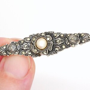 May include: A silver brooch with a pearl center and marcasite accents. The brooch is in the shape of a long, narrow oval.