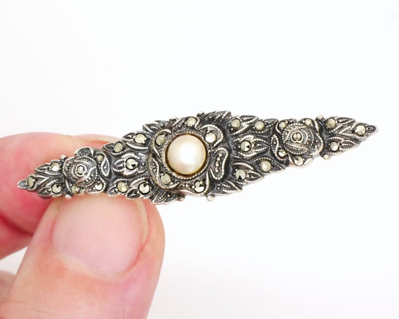 May include: A silver brooch with a pearl center and marcasite accents. The brooch is in the shape of a long, narrow oval.