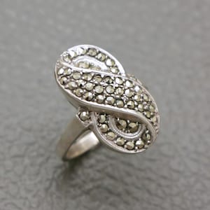 Könnte beinhalten: Silberfarbener Ring mit einem länglichen, ovalen Design, besetzt mit Markasitsteinen. Der Ring hat ein dekoratives, wirbelndes Muster und einen Ring. Die Markasitsteine sind klein und rund.