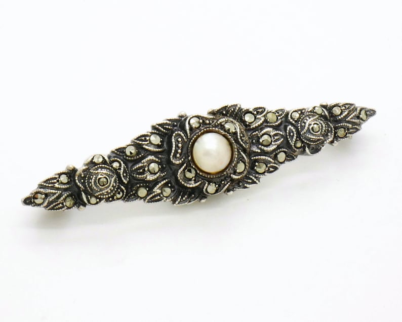 May include: A silver brooch with a pearl center and intricate marcasite detailing. The brooch is shaped like a long, narrow oval.