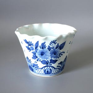 May include: White ceramic planter with a ruffled rim, adorned with hand-painted blue floral designs. The planter features a detailed blue floral pattern around the body and a decorative border near the base. A charming piece for displaying plants.