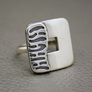 Può includere: Anello d'argento con una faccia quadrata e un quadrato ritagliato. Un lato dell'anello presenta un design strutturato grigio scuro. L'anello ha un'estetica minimalista e moderna.