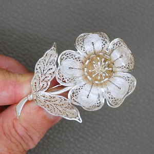 Op de afbeelding: Zilveren filigraan bloembroche met een gedetailleerd open ontwerp. De broche heeft een grote bloem met een centraal, goudkleurig element en een delicaat blad. Het ingewikkelde metaalwerk creëert een kantachtig uiterlijk.