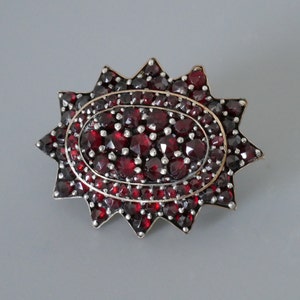 Op de afbeelding: Een vintage broche met een sterrenpatroon. De broche heeft een centrale ovale cluster van dieprode edelstenen, omgeven door een zilverkleurig frame en een stervormige rand. De edelstenen zijn dicht op elkaar geplaatst, wat een rijke, getextureerde uitstraling creëert.