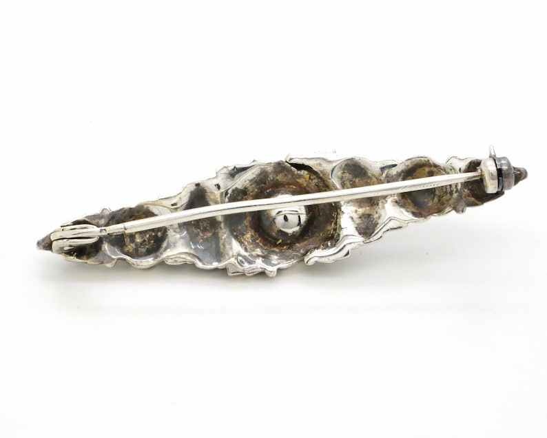 May include: A silver brooch with a detailed, organic design. The brooch is shaped like a leaf and has a textured surface. The brooch has a pin and catch closure.