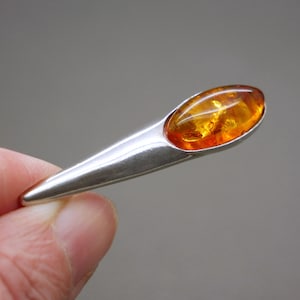 Può includere: Un pendente in argento con una pietra d'ambra a forma di goccia. L'ambra è di un colore arancione caldo e traslucido, e l'incastonatura in argento ha una finitura lucida. Il pendente è tenuto da una mano su uno sfondo neutro.