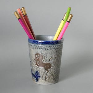 Può includere: Un portapenne in ceramica con un disegno di cavallo, contenente diverse matite colorate. Il portapenne è grigio con un bordo blu e un'illustrazione di cavallo marrone. Le matite sono rosa, rosse, gialle e verdi.