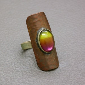 Può includere: Un anello in rame martellato con una grande pietra preziosa di forma ovale. La pietra preziosa è un degradé di rosa, arancio e giallo.