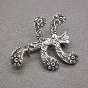 Op de afbeelding: Een zilverkleurige metalen broche met een abstracte, gebogen vorm en gedetailleerde, in reliëf aangebrachte bloem- en krulpatronen. Het ontwerp heeft drie opvallende, afgeronde uiteinden, elk versierd met een cirkelvormig bloemmotief en textuurdetails.