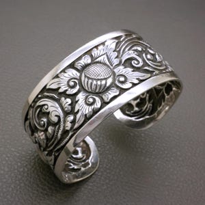 Könnte beinhalten: Ein breiter Silberarmreif mit einem komplexen, geprägten Blumenmuster, einem zentralen Blütenmotiv und geschwungenen Blattdetails. Das Metall hat eine polierte Oberfläche an den erhabenen Stellen und eine dunkle, vertiefte Textur.