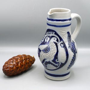 Può includere: Un'anfora in ceramica bianca con un motivo floreale blu e bianco e un uccello. L'anfora ha un manico e un beccuccio.