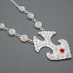 May include: A silver necklace with a large, ornate pendant featuring a stylized cross design. The pendant is decorated with small, round beads and a single red gemstone.