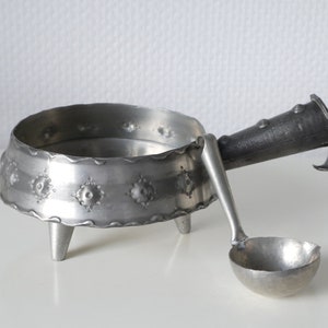 Op de afbeelding: Een zilveren tinnen serviesset met een kleine lepel en een kom met drie poten. De kom heeft een decoratief ontwerp rond de rand.