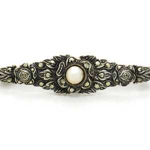 May include: A silver brooch with a pearl center and intricate marcasite detailing. The brooch is shaped like a long oval and has a delicate, ornate design.