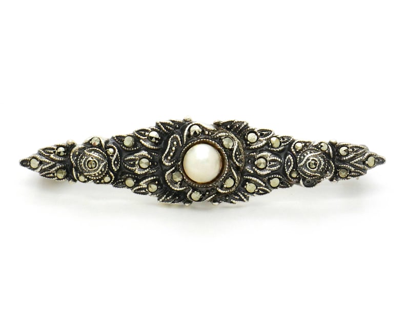May include: A silver brooch with a pearl center and intricate marcasite detailing. The brooch is shaped like a long oval and has a delicate, ornate design.