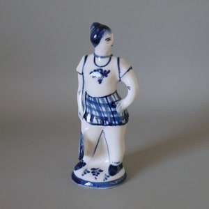 Könnte beinhalten: Eine blaue und weiße Keramikfigur einer Frau, die einen karierten Rock trägt und einen Korb hält. Die Figur ist im traditionellen Volksstil bemalt.