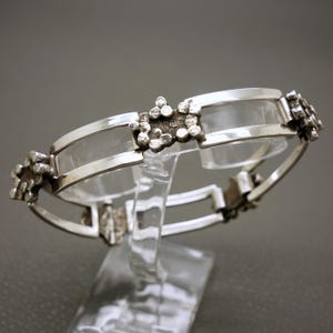 May include: A silver bracelet with a geometric design. The bracelet is made of two bands of silver with a clear glass insert between them. The geometric design is made of small silver squares.