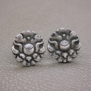 Peut inclure: Une paire de boucles d'oreilles à clous argentées. Chaque boucle d'oreille présente un motif floral détaillé avec un bouton central entouré de pétales superposés. Les boucles d'oreilles ont une esthétique vintage ou antique.