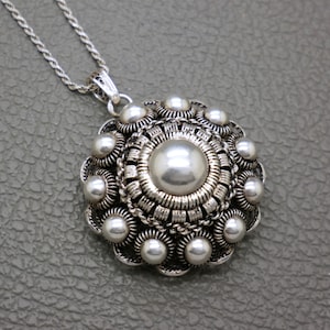 Peut inclure: Collier pendentif argenté avec une grosse perle centrale entourée de petites perles similaires. Le pendentif présente un design détaillé et est suspendu à une chaîne décorative. Le collier est présenté sur une surface grise.
