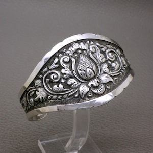 Könnte beinhalten: Ein breiter Armreif aus Silber mit einem detaillierten, geprägten Blumenmotiv und einer zentralen Knospe. Die Metalloberfläche hat polierte Kanten und einen abgedunkelten, vertieften Hintergrund, der die feinen Rankenmuster hervorhebt.