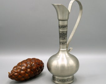 May include: A silver pewter pitcher with a decorative design of grapes and vines. The pitcher has a round body and a long, slender neck with a spout. The handle is curved and attached to the neck.