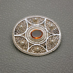 Op de afbeelding: Een zilveren filigraan broche met een ronde, oranje edelsteen in het midden.