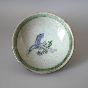 Op de afbeelding: Een kleine, ronde keramische kom met een lichtgroen interieur en een donkerdere groene rand. De kom heeft een blauw en wit hertenontwerp met bloemaccenten. De kom is waarschijnlijk handgemaakt en heeft een rustieke uitstraling.