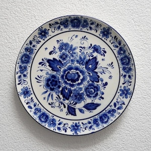 Può includere: Un piatto rotondo in ceramica con base bianca e complessi motivi floreali blu. Il centro mostra un grande bouquet di vari fiori e foglie, mentre il bordo è decorato con una bordura continua di piccoli fiori e viticci.