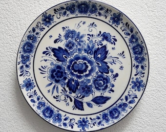 May include: A round ceramic plate featuring a white base with intricate blue floral patterns. The center displays a large bouquet of various flowers and leaves, while the rim is decorated with a continuous border of smaller blossoms and vines.