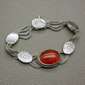 May include: A silver bracelet with a red carnelian stone and intricate floral designs. The bracelet has multiple strands of chain and a clasp.