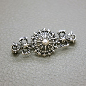 May include: A silver brooch with a central circular design and two smaller bow-shaped designs on either side.