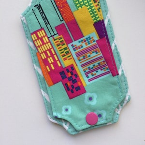 Pode incluir: Uma etiqueta de bagagem de tecido azul turquesa com um design de cidade colorida. A etiqueta tem uma borda branca em ziguezague e um botão rosa.