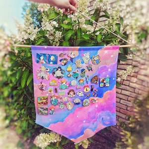 Op de afbeelding: Een kleurrijke banner met een pastelkleurig hemelontwerp, met een verzameling emaille pins. De pins tonen verschillende cartoonkarakters en ontwerpen, bevestigd aan de stof. De banner wordt opgehangen aan een houten stok en touw.