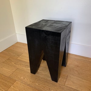May include: A black wooden stool with a square top and a unique, angled leg design. The stool is made of solid wood and has a rustic, natural finish.