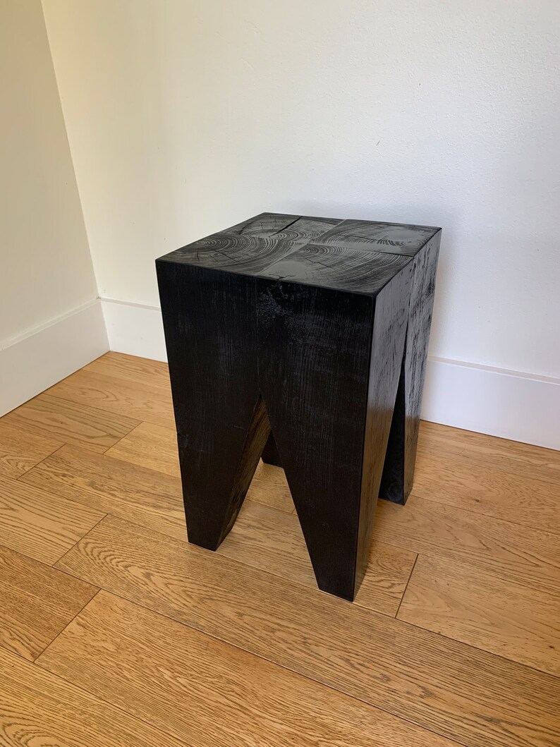 May include: A black wooden stool with a square top and a unique, angled leg design. The stool is made of solid wood and has a rustic, natural finish.