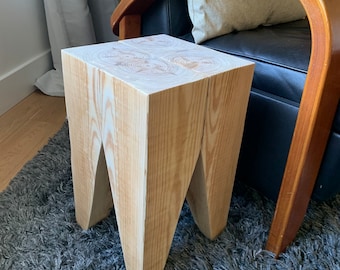 May include: A light wood stool with a unique, angular design. The stool has four legs that are shaped like a V. The stool is sitting on a gray rug.