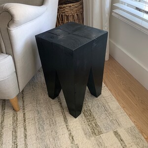 May include: A black wooden stool with a square top and four legs. The stool is sitting on a rug with a light brown and gray pattern.