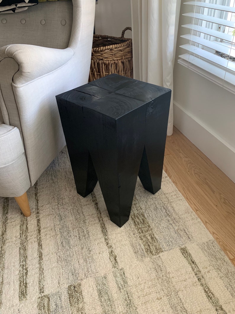 May include: A black wooden stool with a square top and four legs. The stool is sitting on a rug with a light brown and gray pattern.