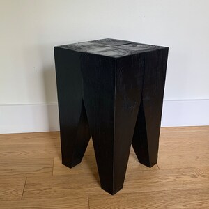 May include: A black wooden stool with a square top and four legs that are angled inwards. The stool is made of solid wood and has a natural finish.