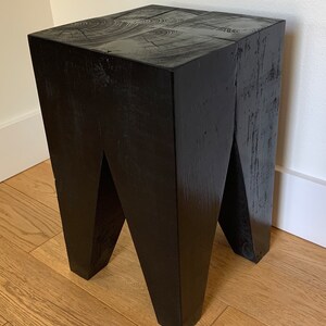 May include: A black wooden stool with a unique, angular design. The stool has a square top and four legs that are angled inwards.