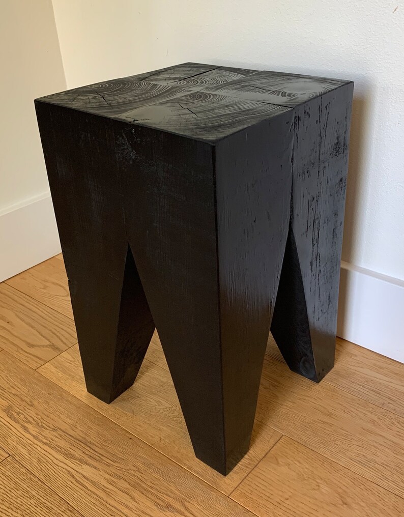 May include: A black wooden stool with a unique, angular design. The stool has a square top and four legs that are angled inwards.