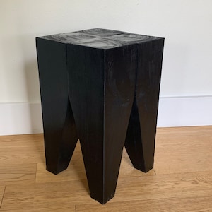 May include: Black wooden stool with a unique, geometric design. The stool has a square top and four legs that are angled inwards.