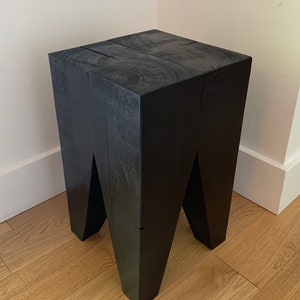 May include: Black wooden stool with a square top and four legs. The legs are angled inwards, creating a unique design.